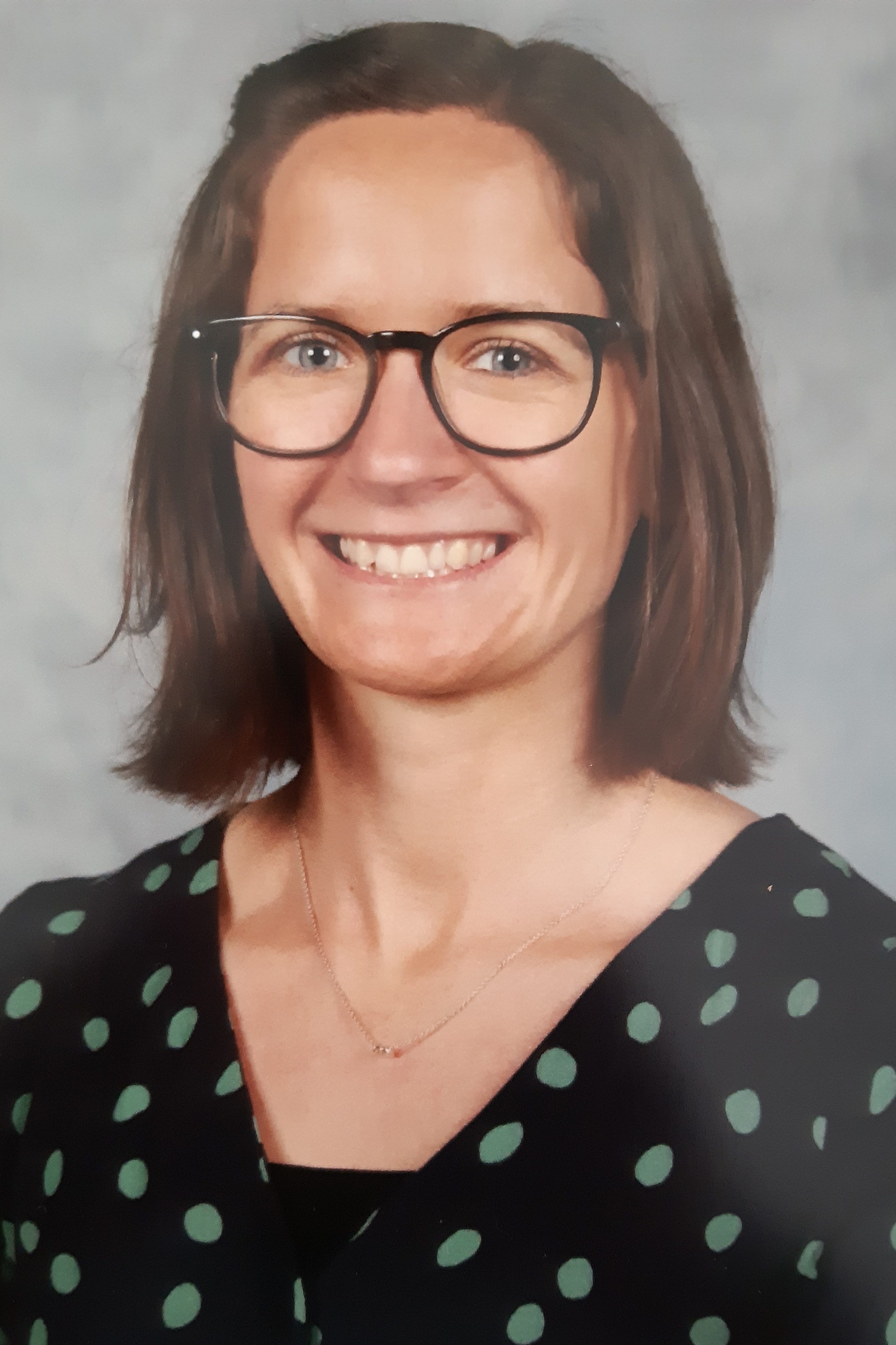 Portrait of a woman with brown hair and glasses, smiling. She is wearing a black top with green polka dots. The image features a light gray background.